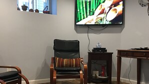 Ein Wohnzimmer mit einem an der Wand befestigten Fernseher, einem Holzstuhl mit gestreifter Sitzkissen, einem kleinen Tisch mit einer Uhr und einem Fenster mit Blick auf Gebäude.