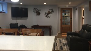 Ein Wohnzimmer mit weißer Arbeitsplatte, einer schwarzen Ledersofa, einem Holzespeisetisch mit Stühlen, einem an der Wand montierten Fernseher und einem Teppich mit Muster.
