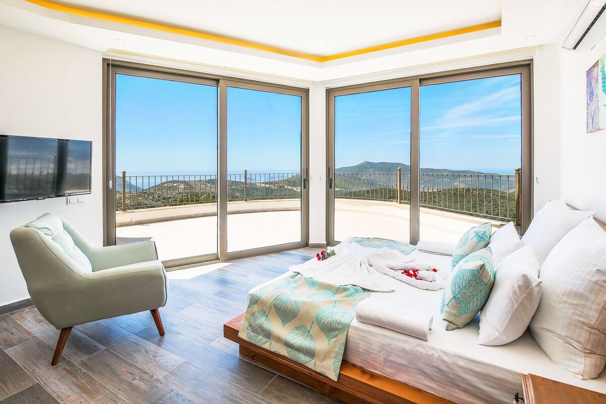 A modern bedroom with a large bed, a chair, and a TV. It has large windows with a view of the sea and hills.