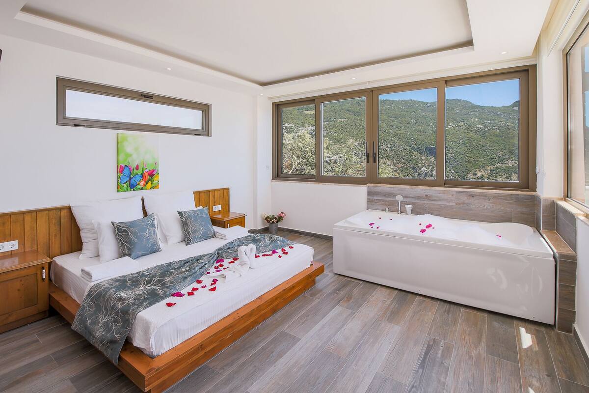 A bedroom with a bed, a wooden headboard, a nightstand, a bedside table, a large window with a view of hills, and a bathtub with rose petals.