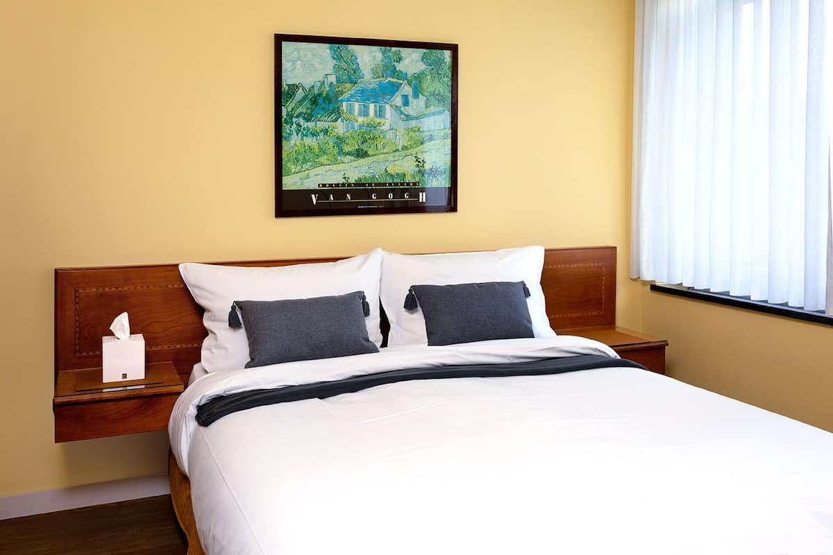 Une chambre d’hôtel avec un lit, deux oreillers, une table de chevet, une boîte de mouchoirs et un tableau représentant une maison accroché au mur.