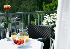 Une table avec une carafe de jus d’orange, un bol de fraises et un verre de jus d’orange sur un balcon, où se trouvent une chaise en osier et une rose dans un vase.