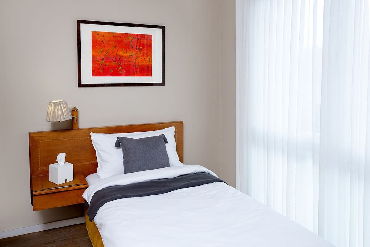 Une chambre d’hôtel avec une tête de lit en bois, une table de chevet avec une lampe, un tableau abstrait encadré et une fenêtre avec des rideaux légers.