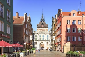 A central building with an ornate facade and a large archway, flanked by colorful buildings and outdoor seating areas with umbrellas.