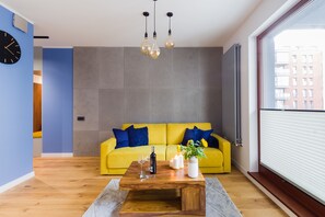 A modern living room with a yellow sofa, a wooden coffee table, and a view of buildings through a window.