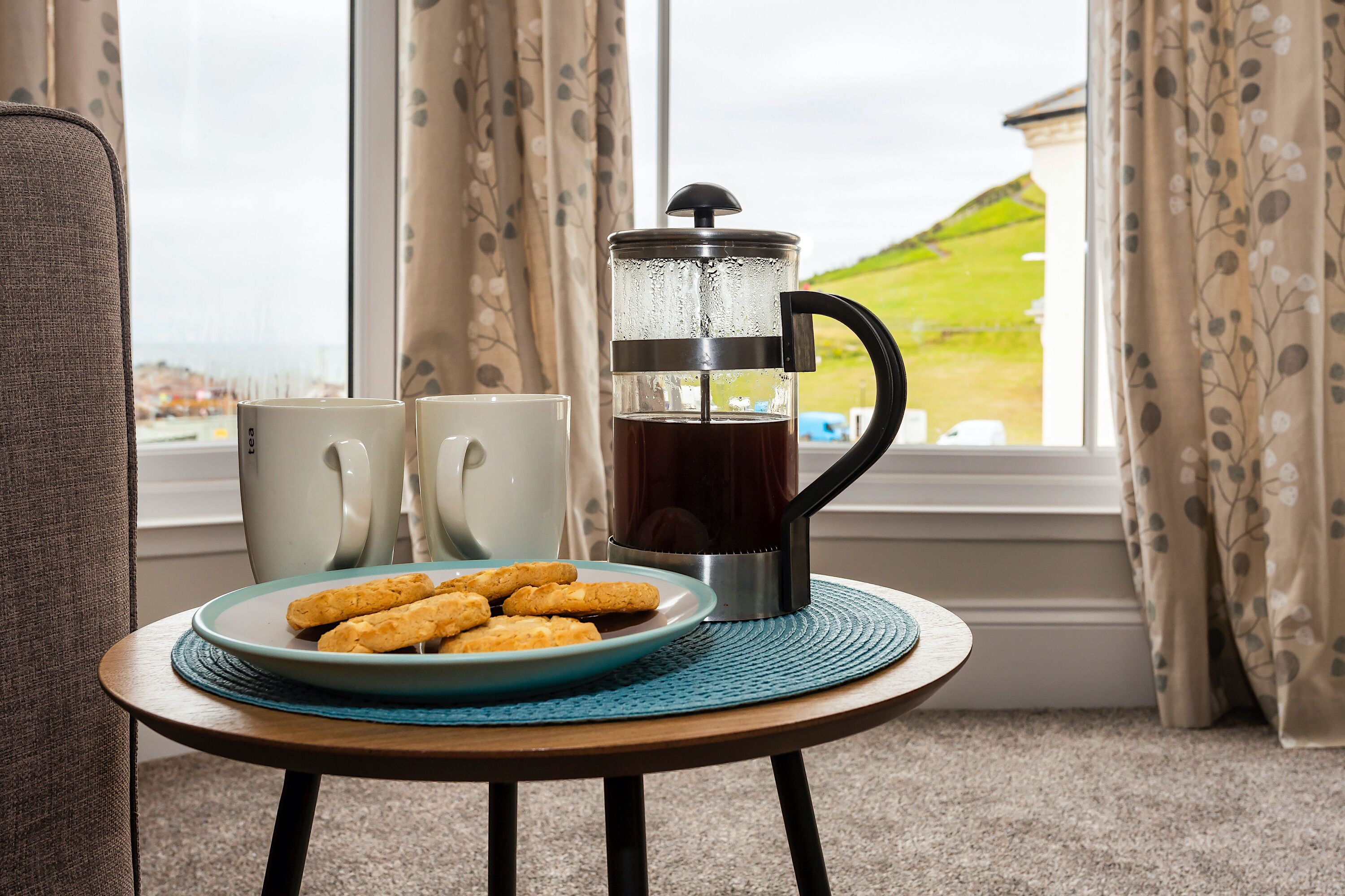 Quarto aconchegante com vista para uma paisagem verde através de uma janela, uma cafeteira, duas canecas brancas e um prato de biscoitos sobre uma mesa redonda pequena.