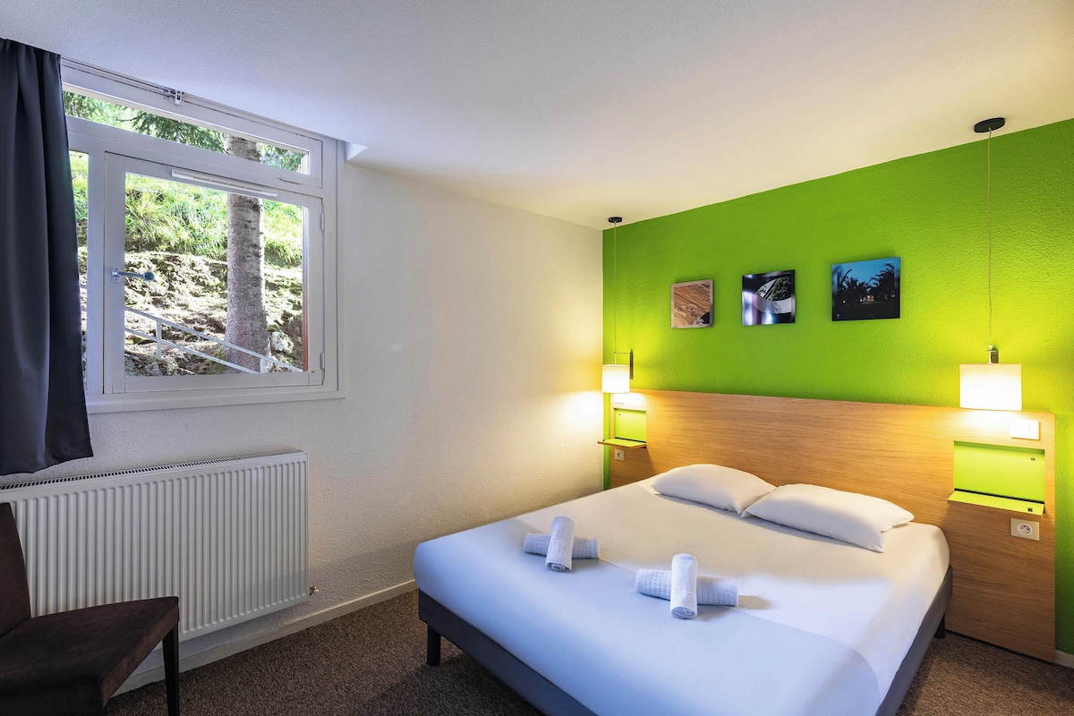 Ein Hotelzimmer mit einem Bett, zwei Nachttischlampen, einem Fenster mit Vorhängen, einem Heizkörper und einer grünen Akzentwand mit zwei Bildern.