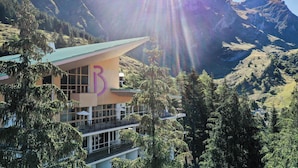 A modern building with a large 'B' on its facade, surrounded by lush greenery and mountains in the background. - Belambra Clubs Gourette - Lou Sarri (Eaux-Bonnes)