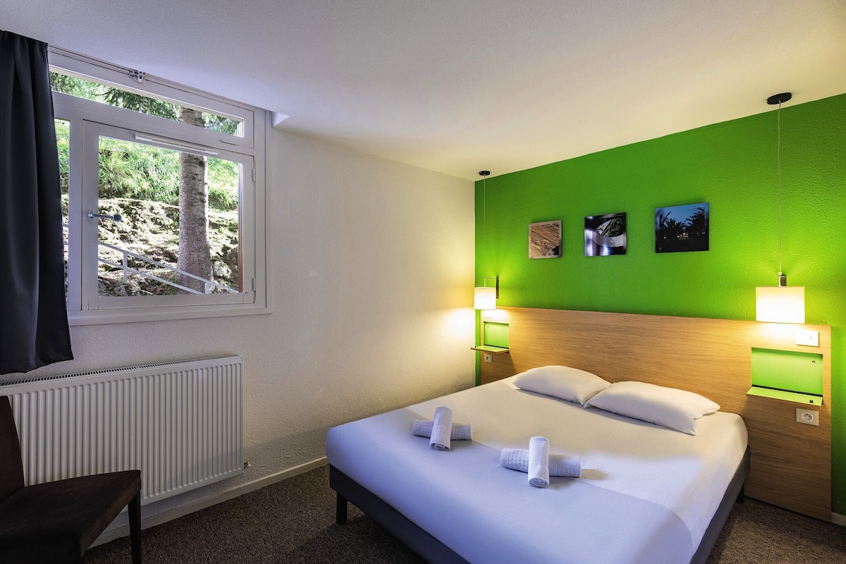 Een hotelkamer met een bed, twee bedlampen, een raam met uitzicht op bomen en twee aan de muur hangende schilderijen.