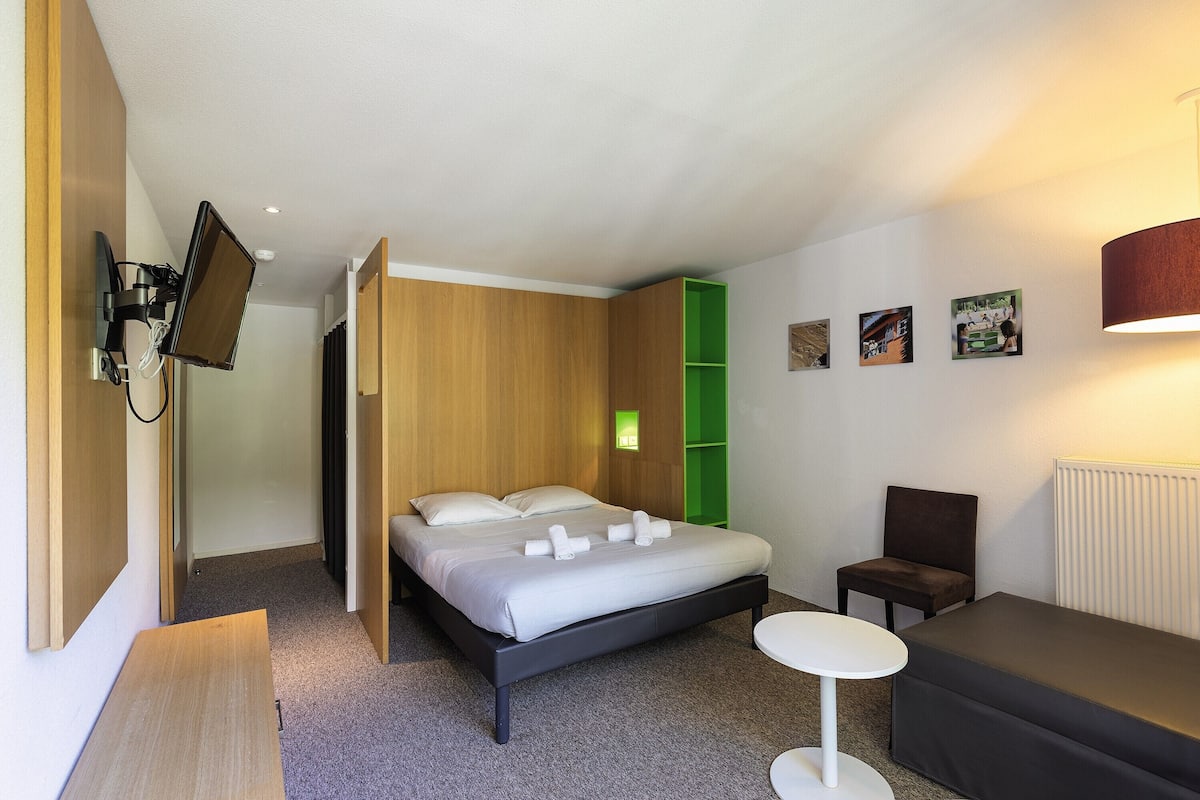 Een moderne hotelkamer met een bed, een klein tafeltje, een stoel en een aan de muur bevestigde televisie.