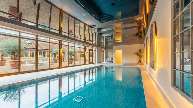 En indendørs swimmingpool med glasvægge, flisegulv og udsigt udad.