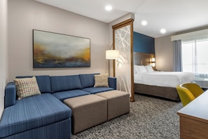 A hotel room with a sectional sofa, a bed, a desk, and a large window.