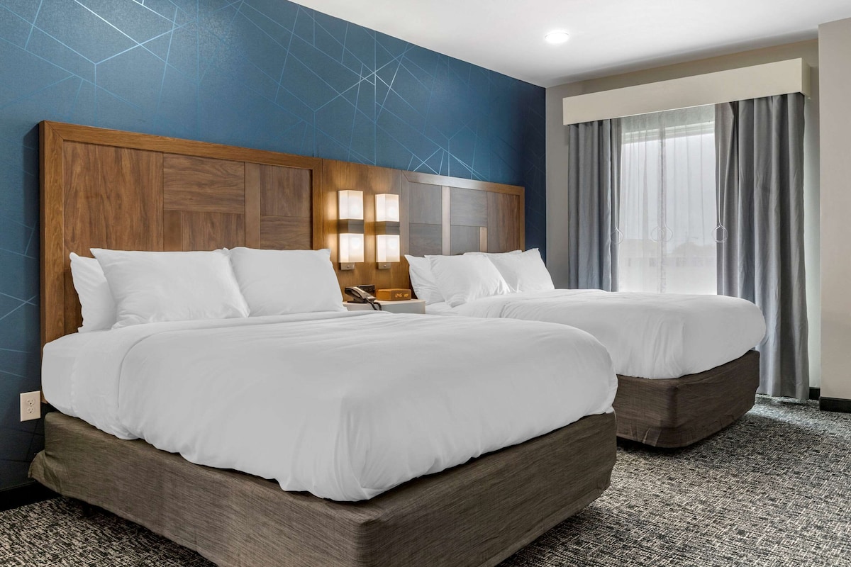 Une chambre d’hôtel avec deux lits, une tête de lit en bois, une grande fenêtre avec des rideaux et une moquette à motifs.
