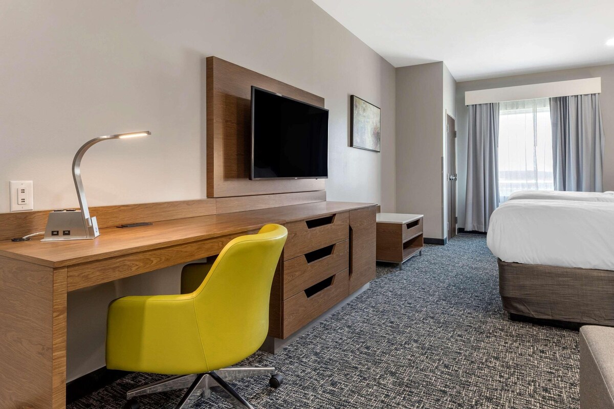 Une chambre d’hôtel avec un bureau en bois, une chaise jaune, un téléviseur à écran plat, un lit et une grande fenêtre avec des rideaux.