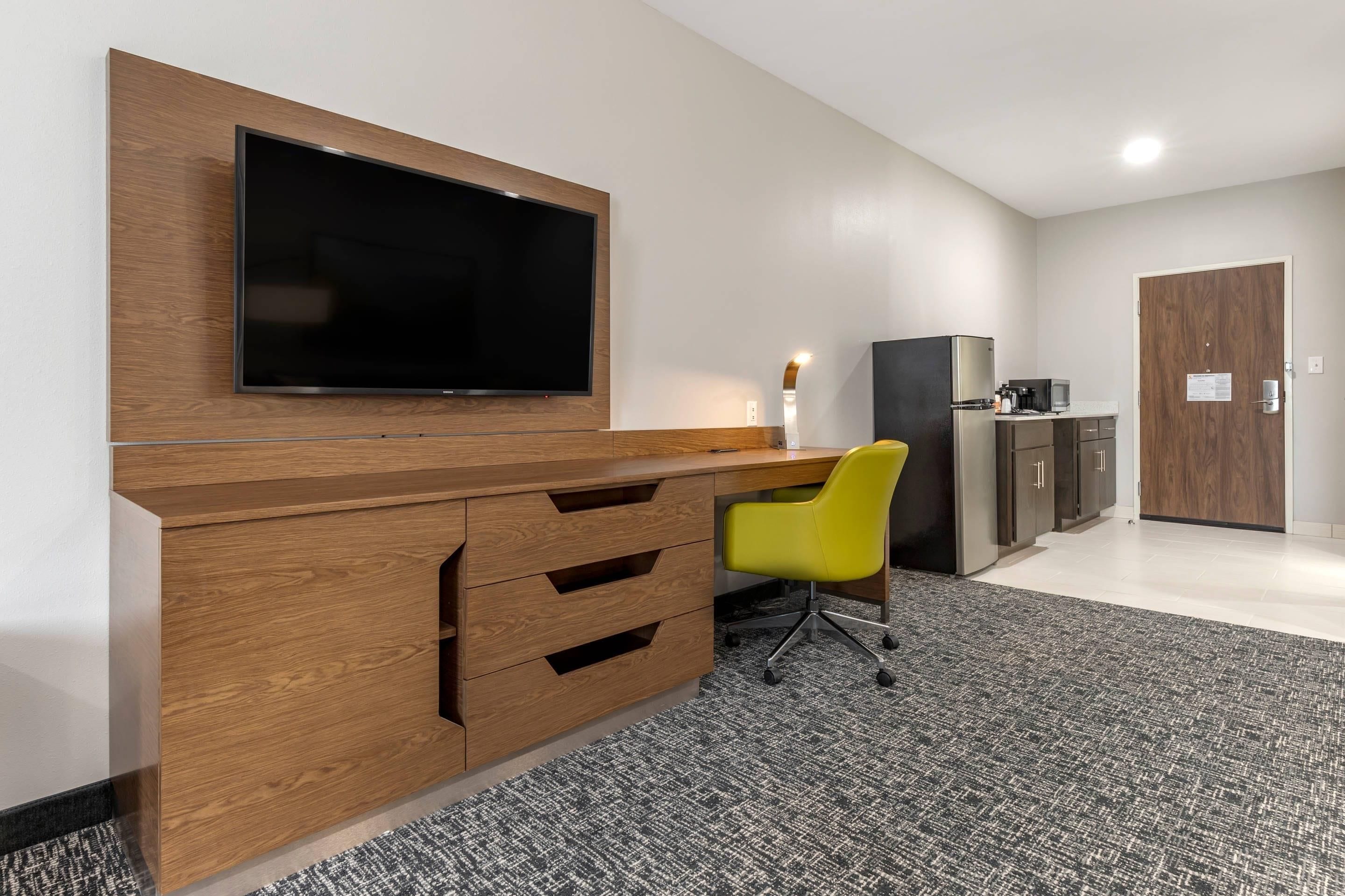 A modern office space with a large flat-screen TV mounted on a wooden desk, a yellow office chair, a refrigerator, and a microwave.
