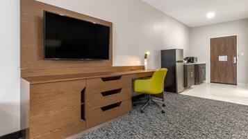A modern office space with a large flat-screen TV mounted on a wooden desk, a yellow office chair, a refrigerator, and a microwave.