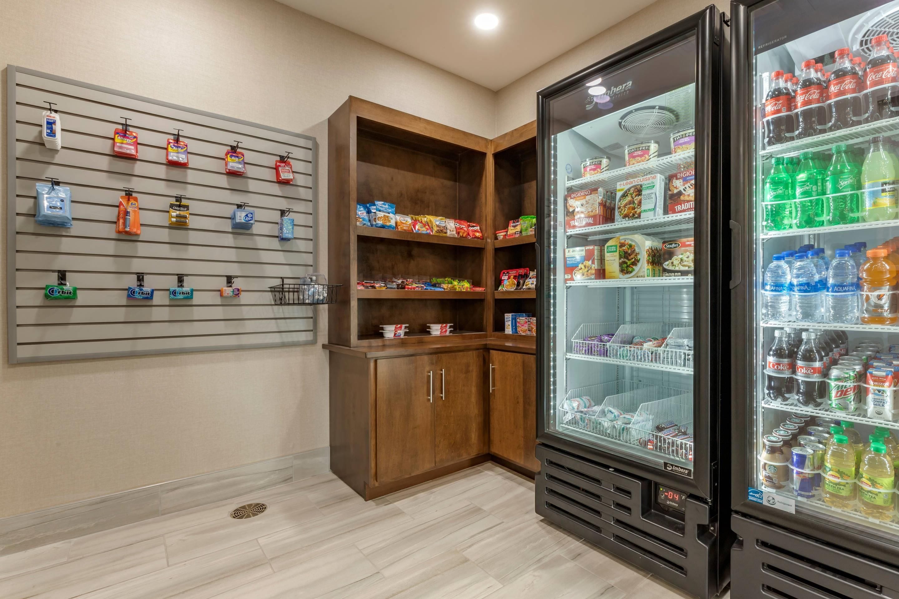 Habitación de hotel con minibar, refrigerador lleno de bebidas y estante con aperitivos y artículos de aseo.