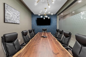 Un salón de conferencias con una larga mesa de madera, sillas de cuero negro, un televisor de pantalla plana montado y una lámpara de araña moderna.