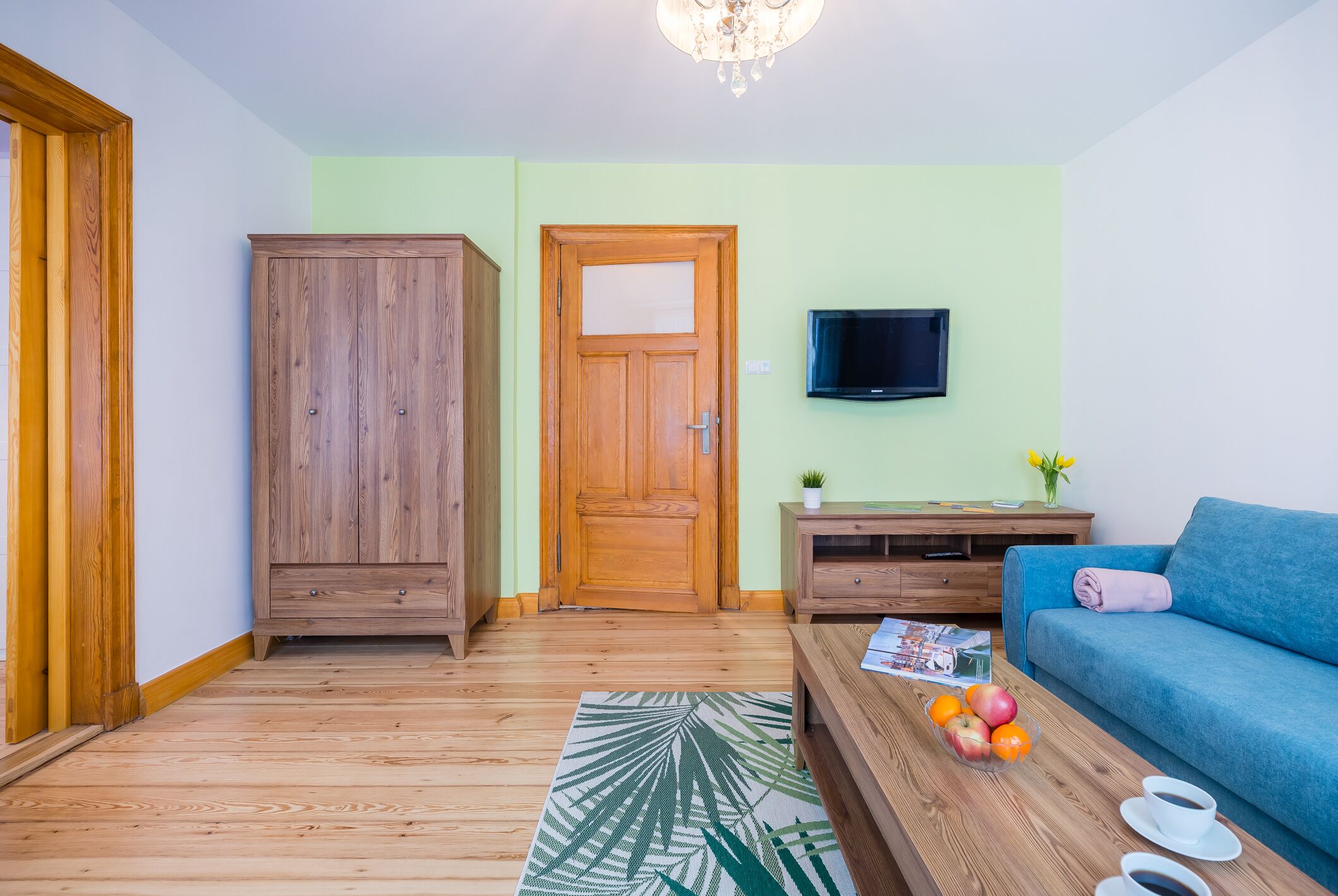 A living room with a wooden wardrobe, a television on the wall, a blue sofa, and a coffee table with fruits and cups.