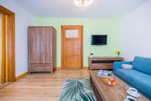 A living room with a wooden wardrobe, a television on the wall, a blue sofa, and a coffee table with fruits and cups. - Freedom Gdansk Apartment (Gdansk)