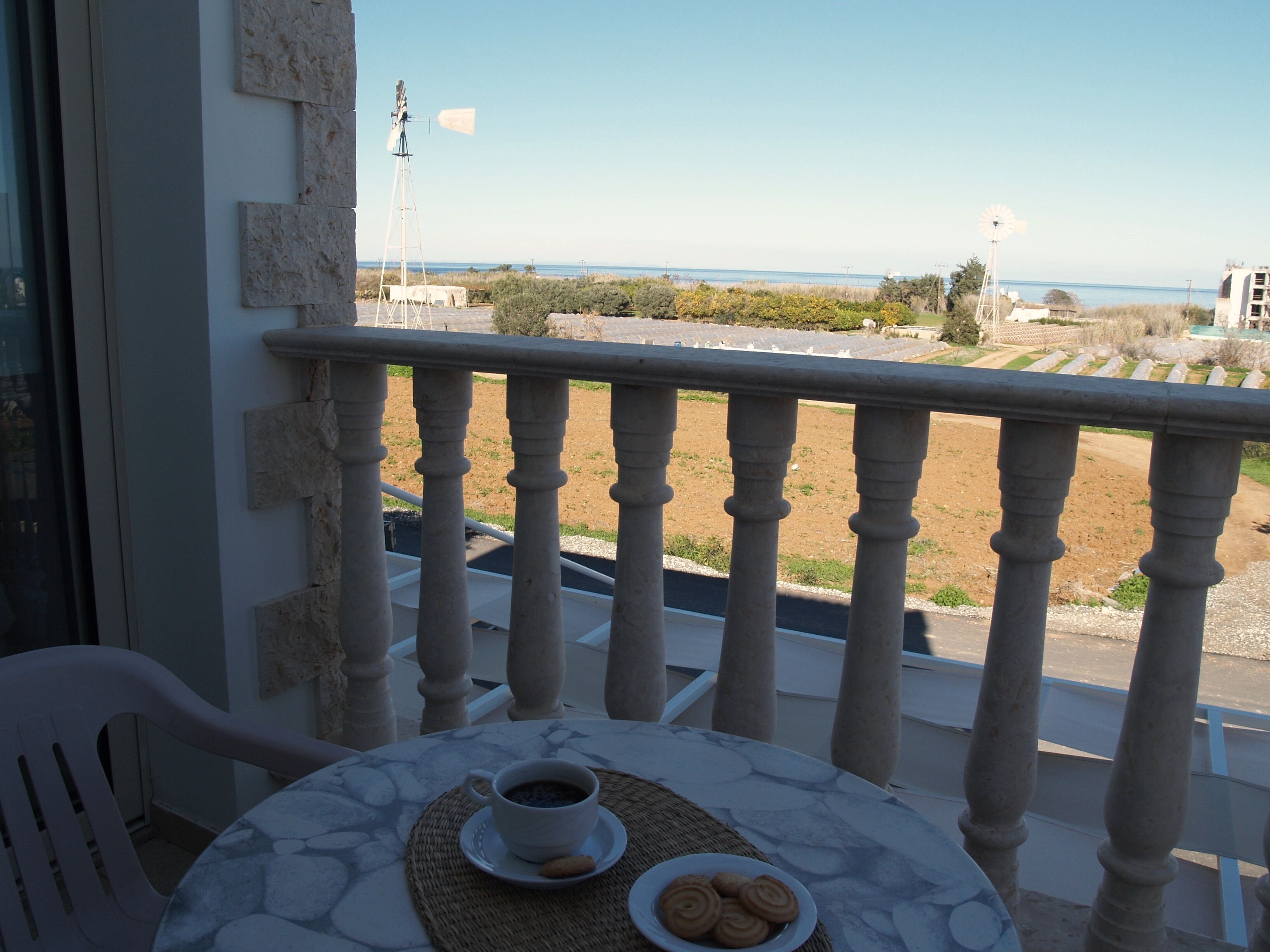 A balcony with a table set for two, a cup of coffee, and a plate of cookies. The view includes a windmill and a beach in the distance.