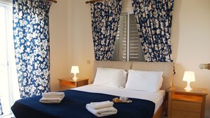 A neatly made bed with white pillows and a blue bedspread, flanked by two bedside tables with lamps, in a room with patterned curtains and a window with blinds. - Villa Ithaca Protaras (Paralimni)