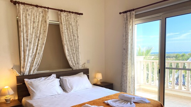 A hotel room with a bed, two pillows, a nightstand with a lamp, and a view of a balcony with a railing. - Villa Ithaca Protaras (Paralimni)