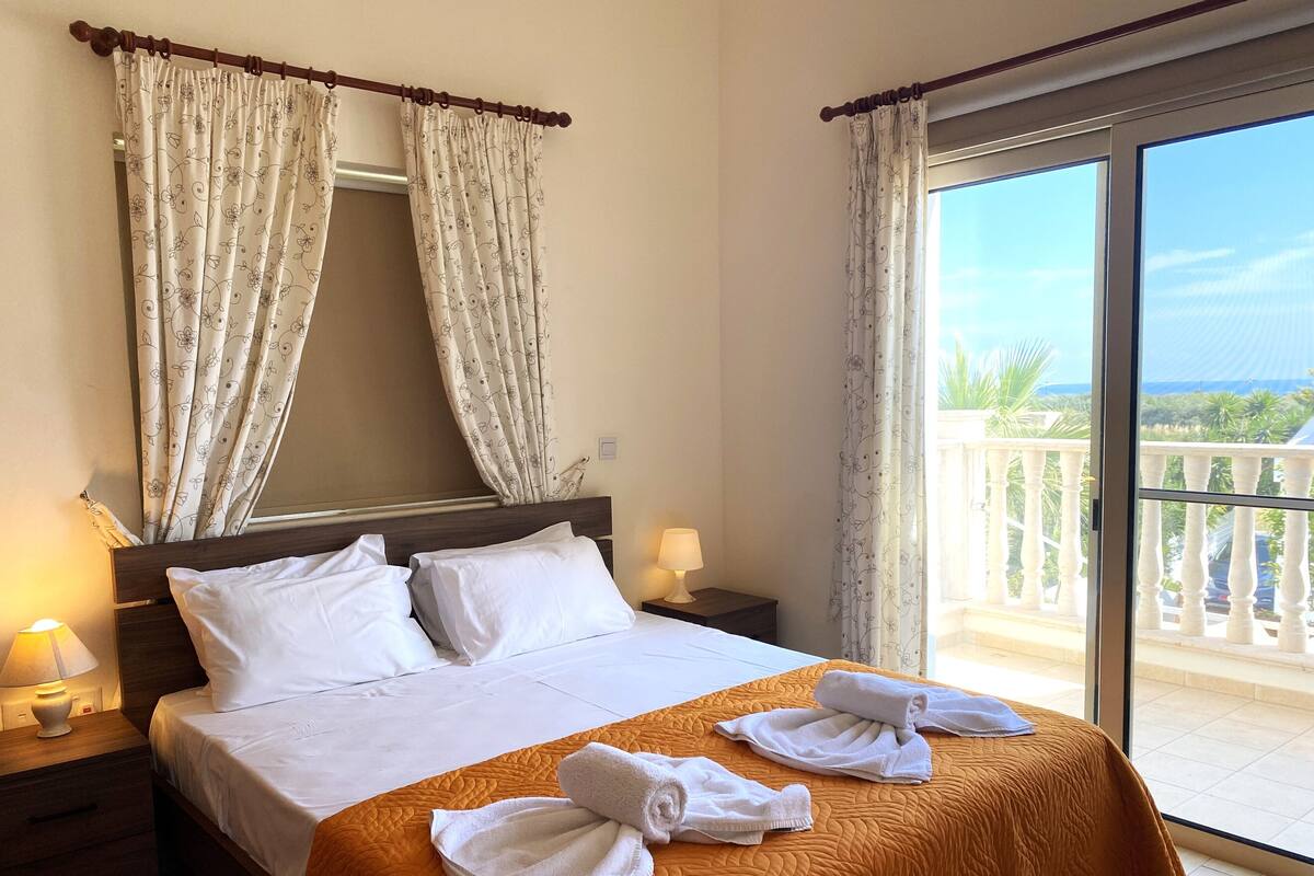 A hotel room with a bed, two pillows, a nightstand with a lamp, and a view of a balcony with a railing.