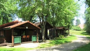A rustic cabin-style lodging with a covered porch and a small deck, surrounded by lush greenery and a clear path leading to the entrance.