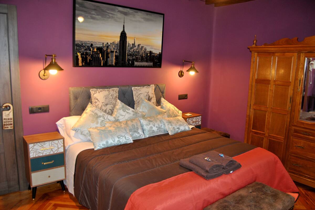 Quarto com cama, duas mesas de cabeceira, um criado-mudo, um guarda-roupa de madeira e uma pintura de paisagem urbana na parede.