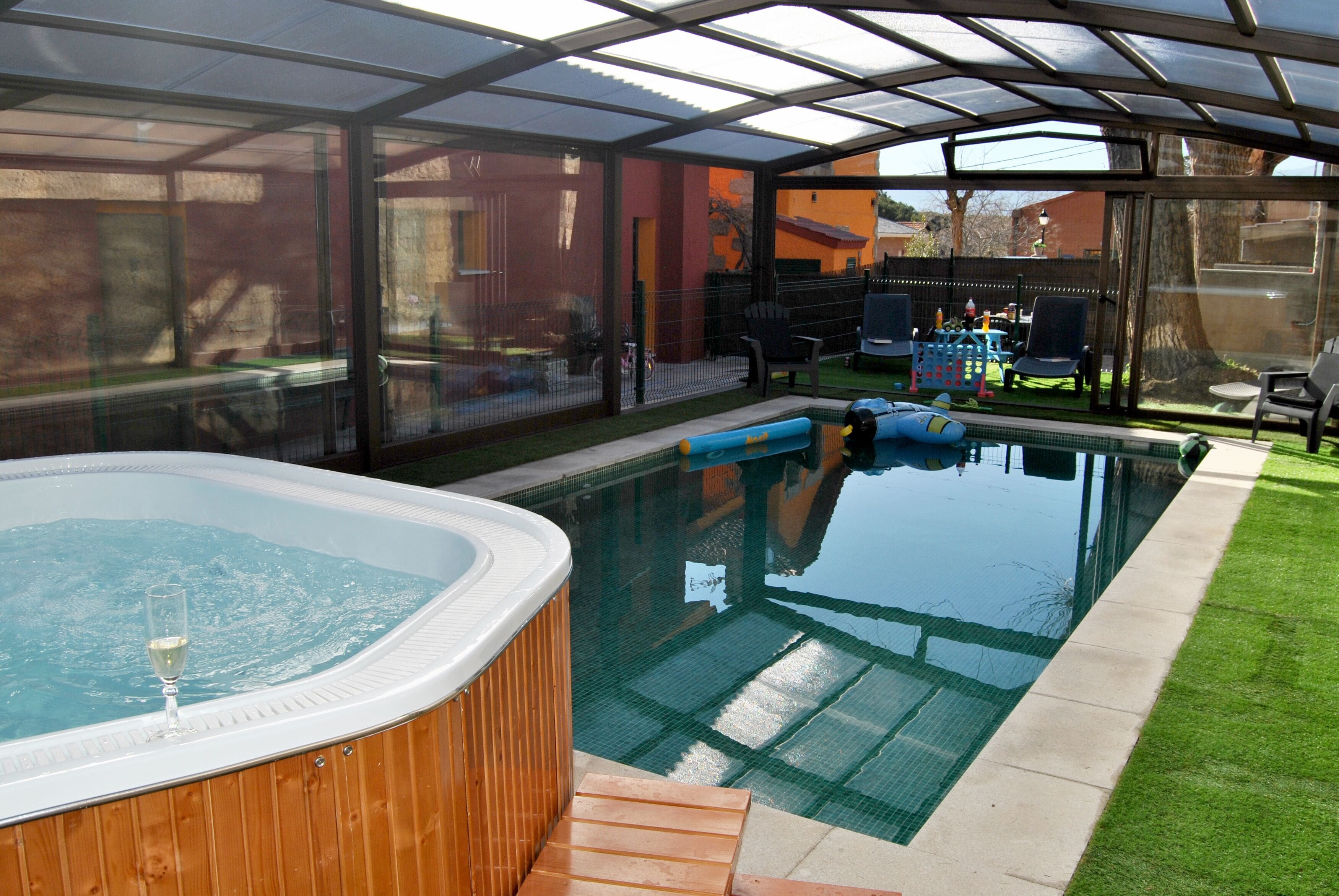 Piscina coberta amb un banyera d'hidromassatge, envoltada per una estructura de vidre i una zona enjardinada amb mobiliari exterior.