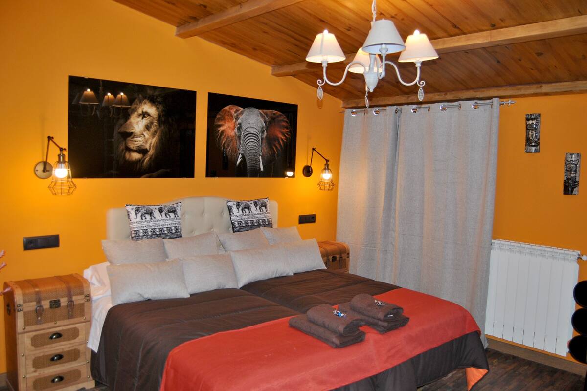 Quarto com uma cama grande, duas luminárias fixadas na parede, teto de madeira e dois retratos emoldurados de animais.