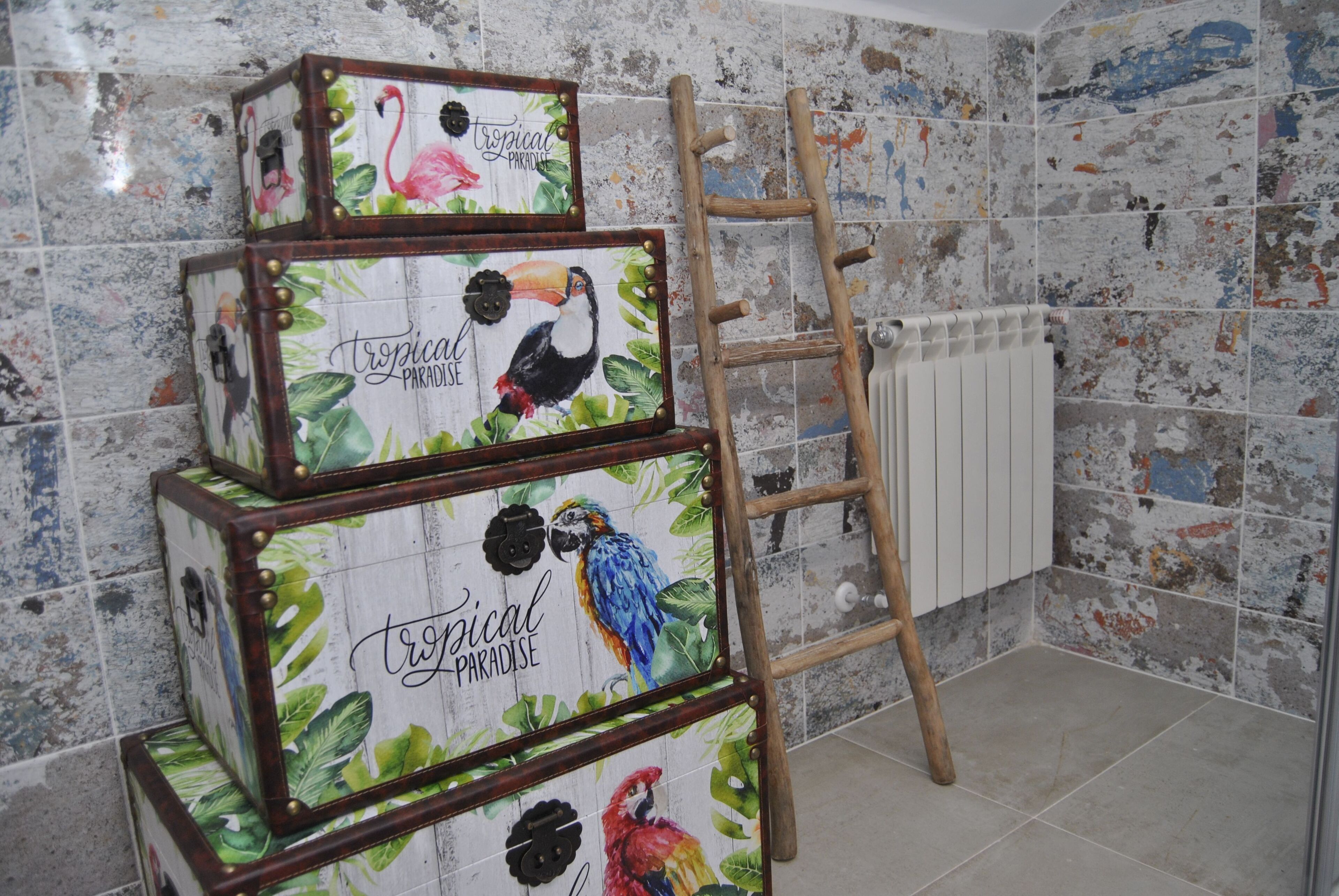 A set of three stacked suitcases with tropical-themed artwork, a wooden ladder, and a radiator on a tiled wall.