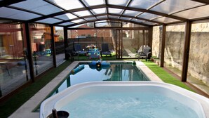 Ein überdachter Pool mit Whirlpool, umgeben von einem eingezäunten Garten mit Liegestühlen und einem Tisch.