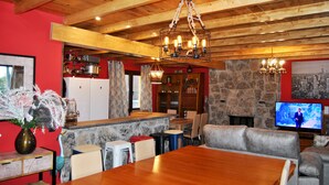 Ein gemütliches Interieur mit Holzbalkendecke, Steinwänden, einem Essbereich und einem Fernseher, der eine Nachrichtensendung zeigt.