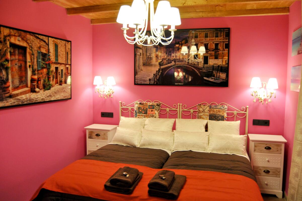Quarto com tema rosa, com uma cama de casal, duas mesas de cabeceira, um candelabro e duas luminárias de parede.