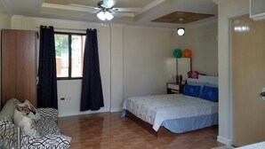 Ein Schlafzimmer mit einem Bett, einem Deckenventilator, einem Fenster mit Vorhängen, einem Holzschrank und einem kleinen Tisch mit Dekoration.