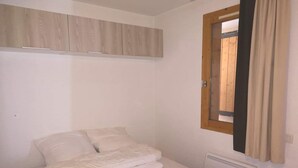 Un lit simple avec une literie blanche, dans une pièce dotée d’une tête de lit en bois, d’une fenêtre avec des rideaux et d’un radiateur.