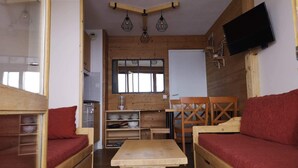 Une pièce confortable, aménagée comme un chalet, avec des murs en bois, une table en bois, deux bancs en bois dotés de coussins rouges, un petit coin cuisine et un téléviseur fixé au mur.