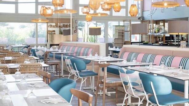 Un restaurant avec de hauts plafonds, des luminaires suspendus en osier et un mélange de chaises bleues et en bois. Les tables sont dressées avec des nappes blanches et de la verrerie élégante.