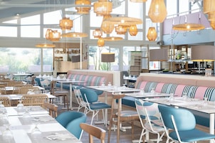 Un restaurante con techos altos, iluminación colgante de mimbre y una mezcla de sillas azules y de madera. Las mesas están dispuestas con manteles blancos y cristalería.