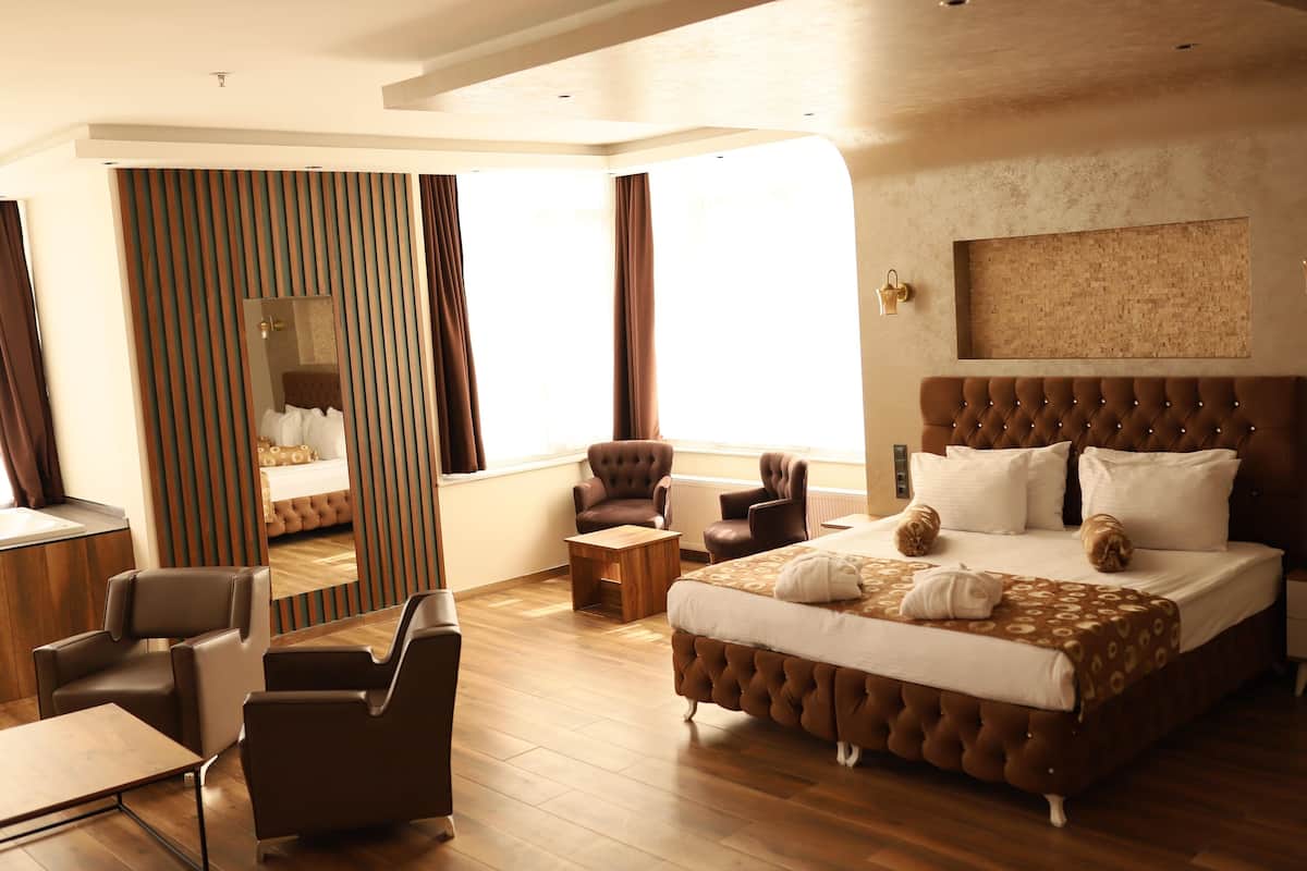A modern hotel room with a large bed, two armchairs, a wooden coffee table, a small side table, and a mirror.