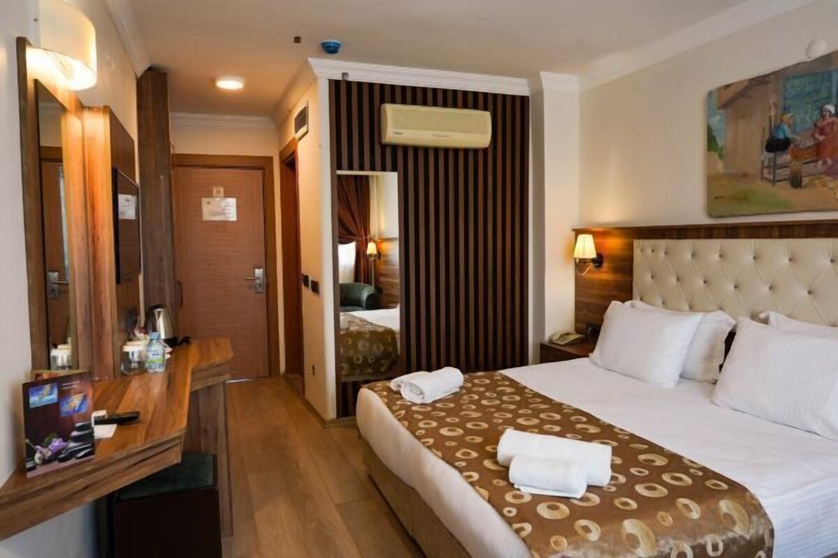 A hotel room with a bed, a wooden desk, a mirror, an air conditioning unit, and a painting on the wall.