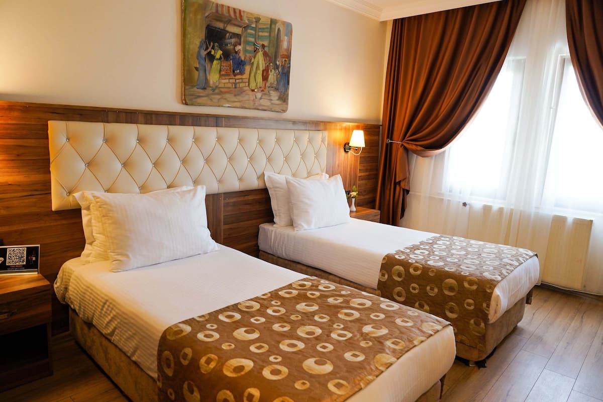 A hotel room with two beds, a wooden headboard, a painting above the beds, a nightstand with a lamp, and a window with curtains.