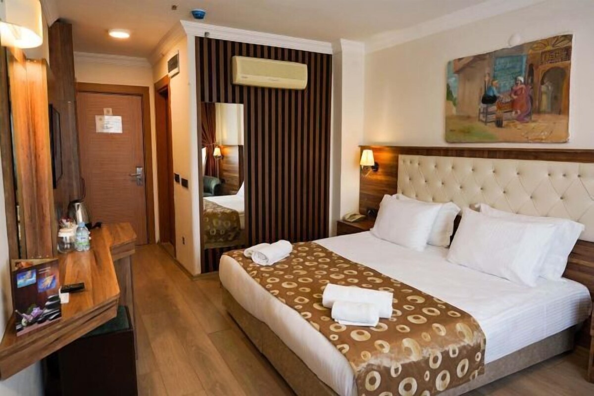 A hotel room with a bed, a desk with a computer, a mirror, a wall-mounted air conditioner, and a painting.