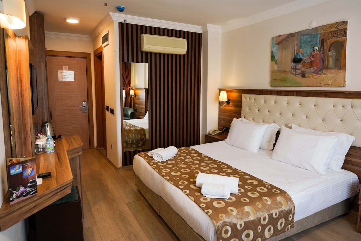 A hotel room with a large bed, a wooden desk, a mirror, a wall-mounted air conditioner, and a painting.