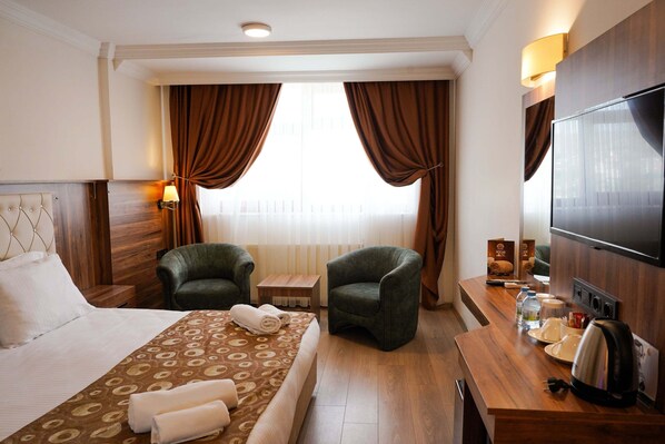 A hotel room with a bed, two armchairs, a wooden desk with a TV, a kettle, and a window with curtains. - Emin Koçak Thermal & Spa - Convention Center (Ürgüp)