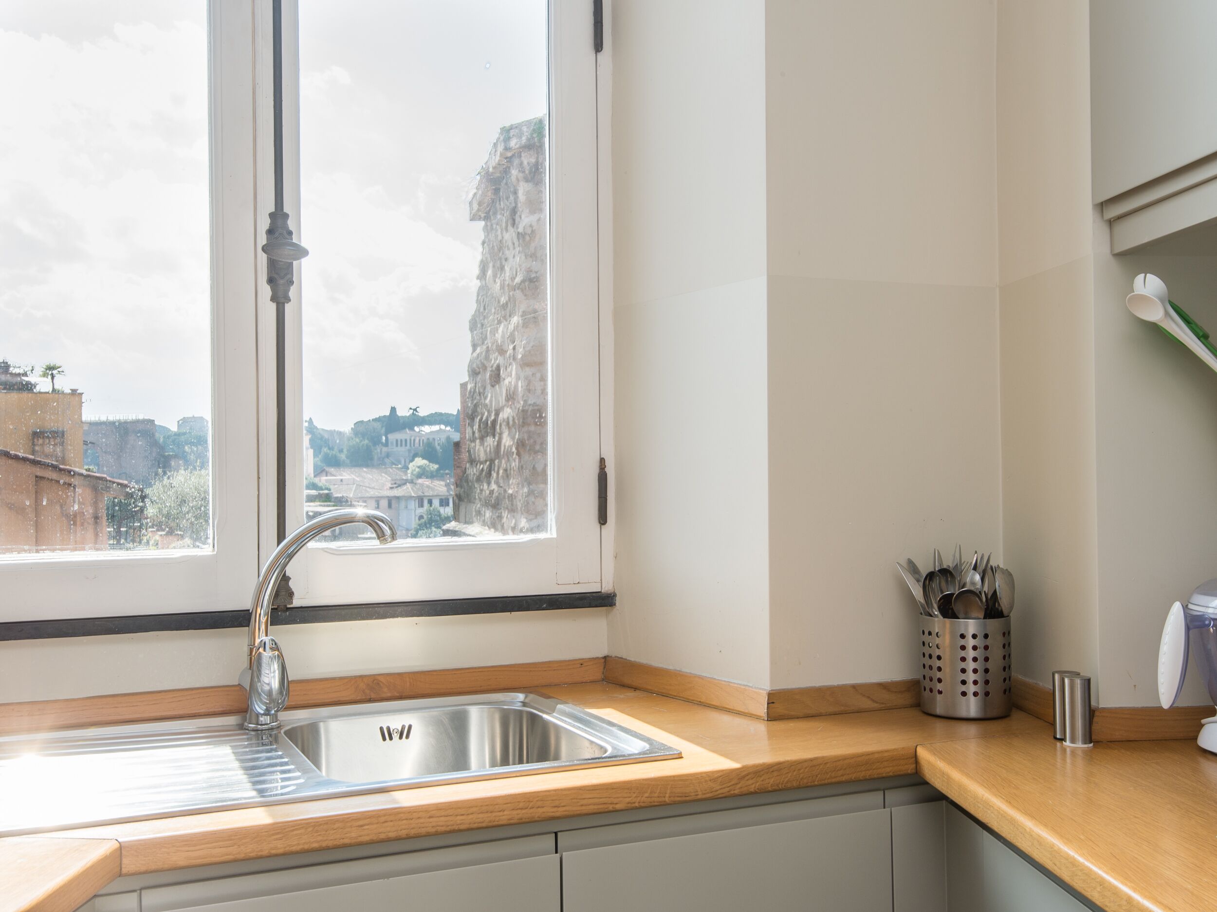 A kitchen with a stainless steel sink, wooden countertop, and a window with a view of buildings and a stone wall.