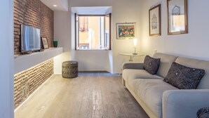 A cozy living room with a brick wall, a wooden ceiling, a sofa with patterned cushions, a TV, and a window with a view. - RSH Luxury One Bedroom Fori Imperiali (Rome)