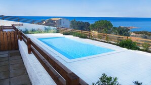 A rectangular swimming pool with a wooden deck and railing, surrounded by a white tiled area, overlooking a coastal landscape with a clear blue sea and distant buildings. - Affittimoderni Isola Rossa Élite - Pati19 (Trinità d'Agultu e Vignola)
